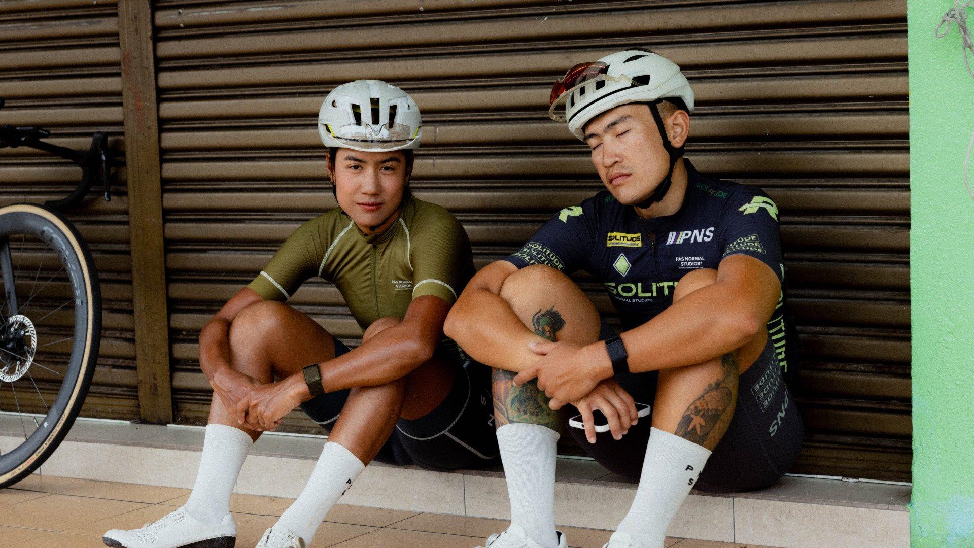 Two cyclists in helmets sitting together, one wearing olive green jersey, other in navy team kit with sponsor logos