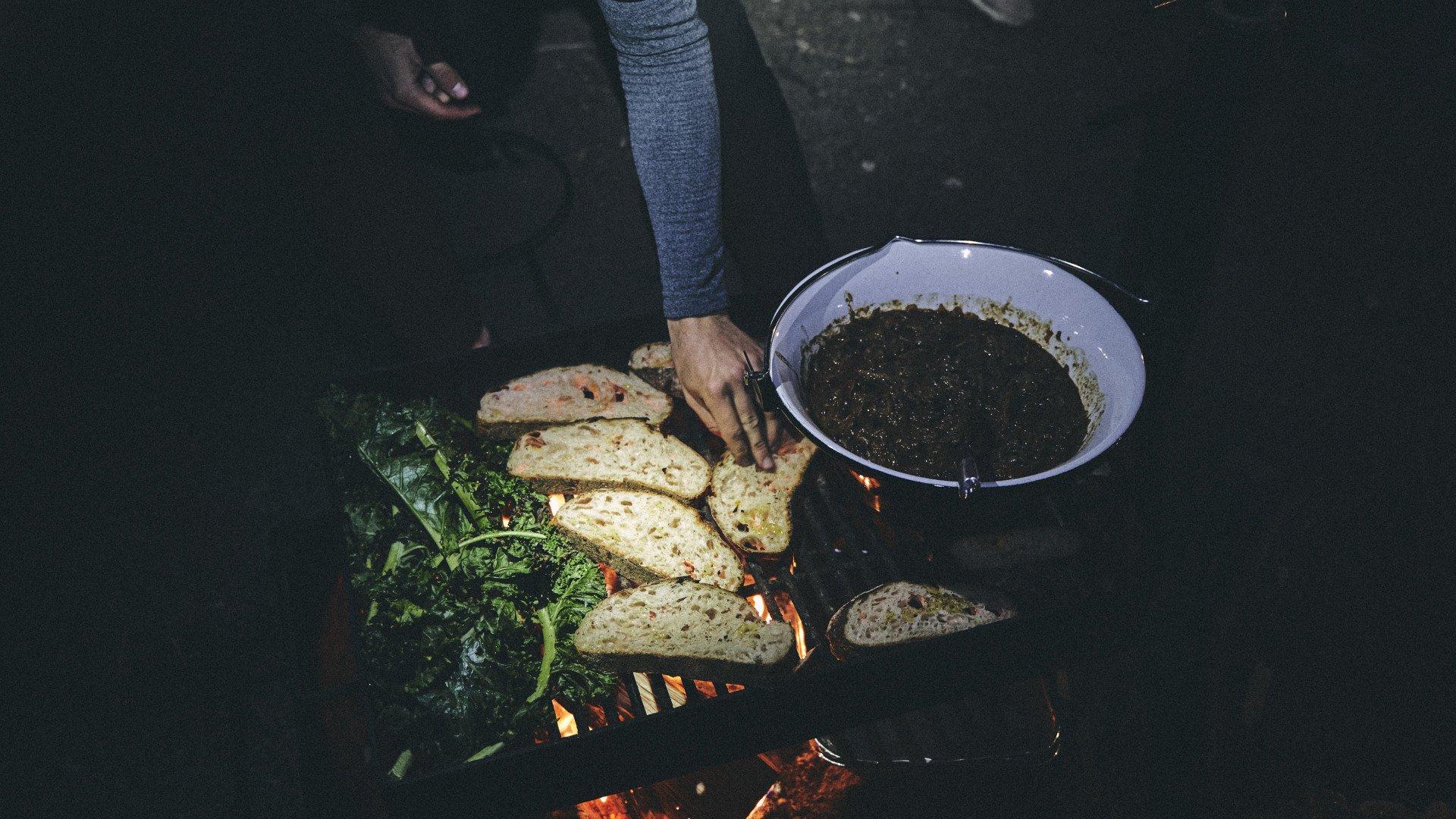 Person cooking bread and greens over campfire with metal pan