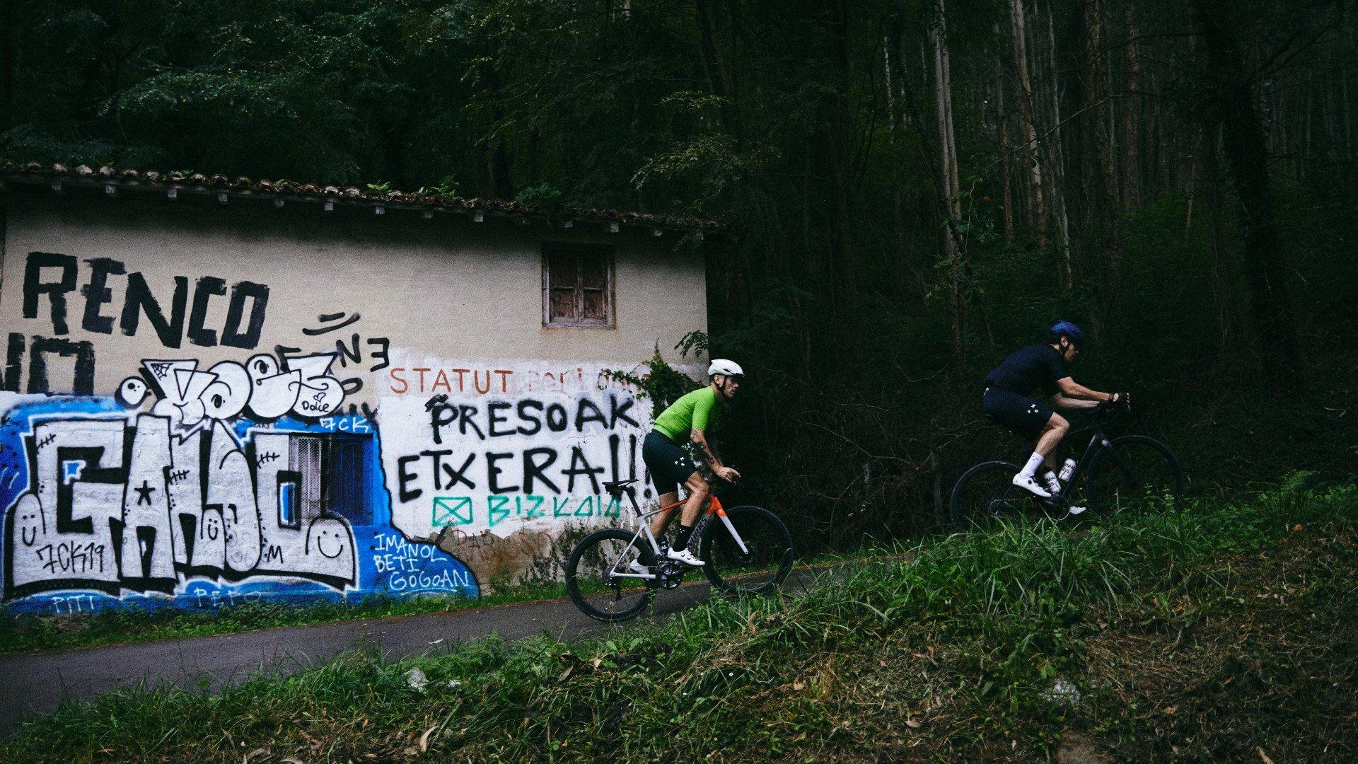 Two cyclists riding past graffitied wall in urban setting, one in green jersey, one in dark blue