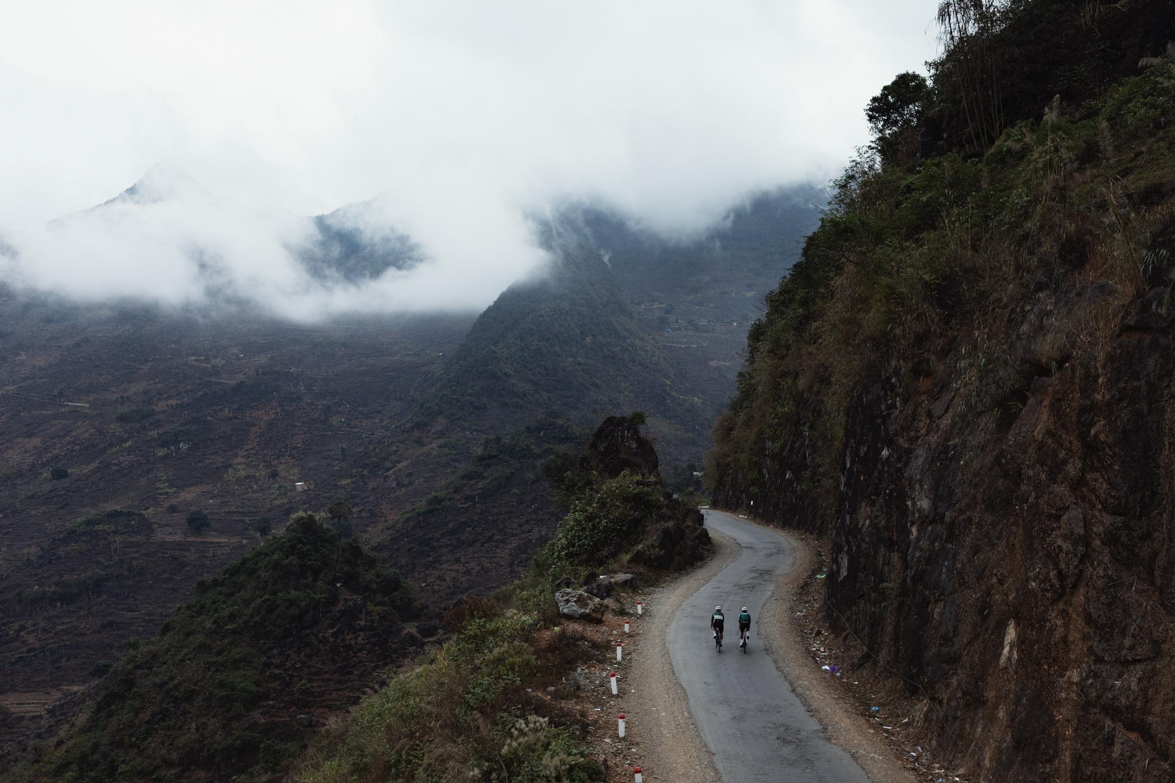 Two cyclists riding through misty mountain valley on winding road with dramatic cloudy landscape