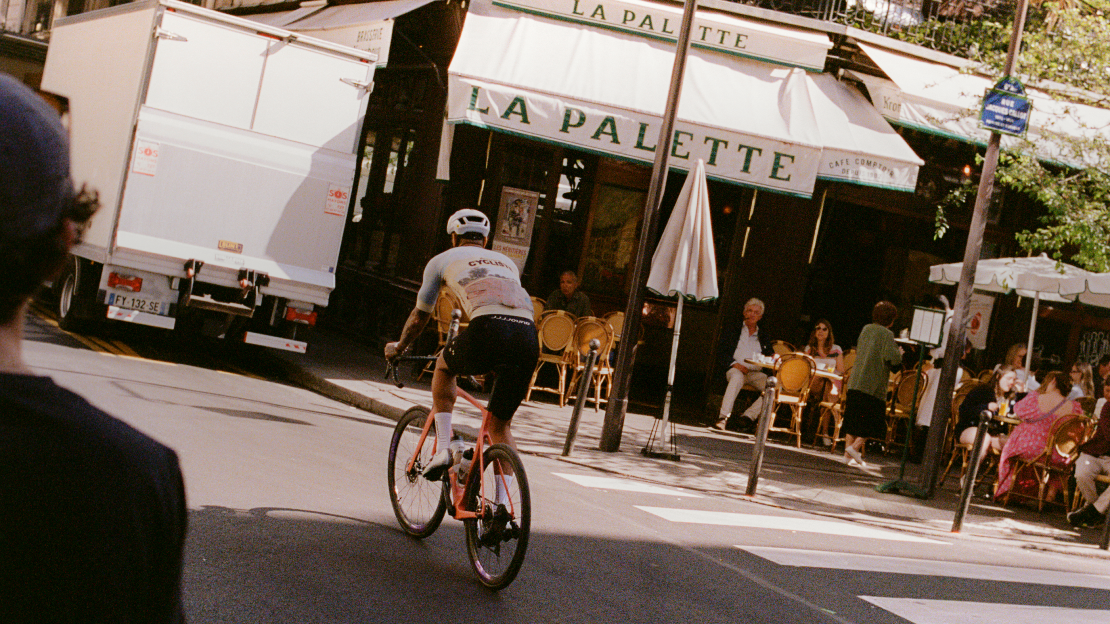 Model wearing white cycling jersey and black shorts cycling past French café