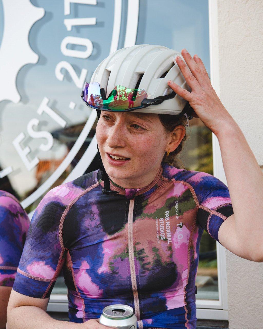 Model wearing multicolored abstract print cycling jersey with white helmet, hand raised to head