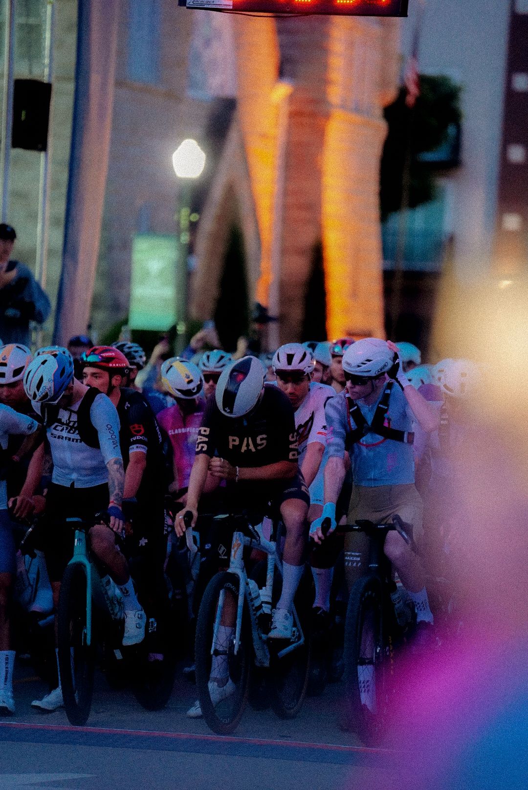 Cyclists in black jersey with chest lettering at start line of gravel race with colorful evening lighting