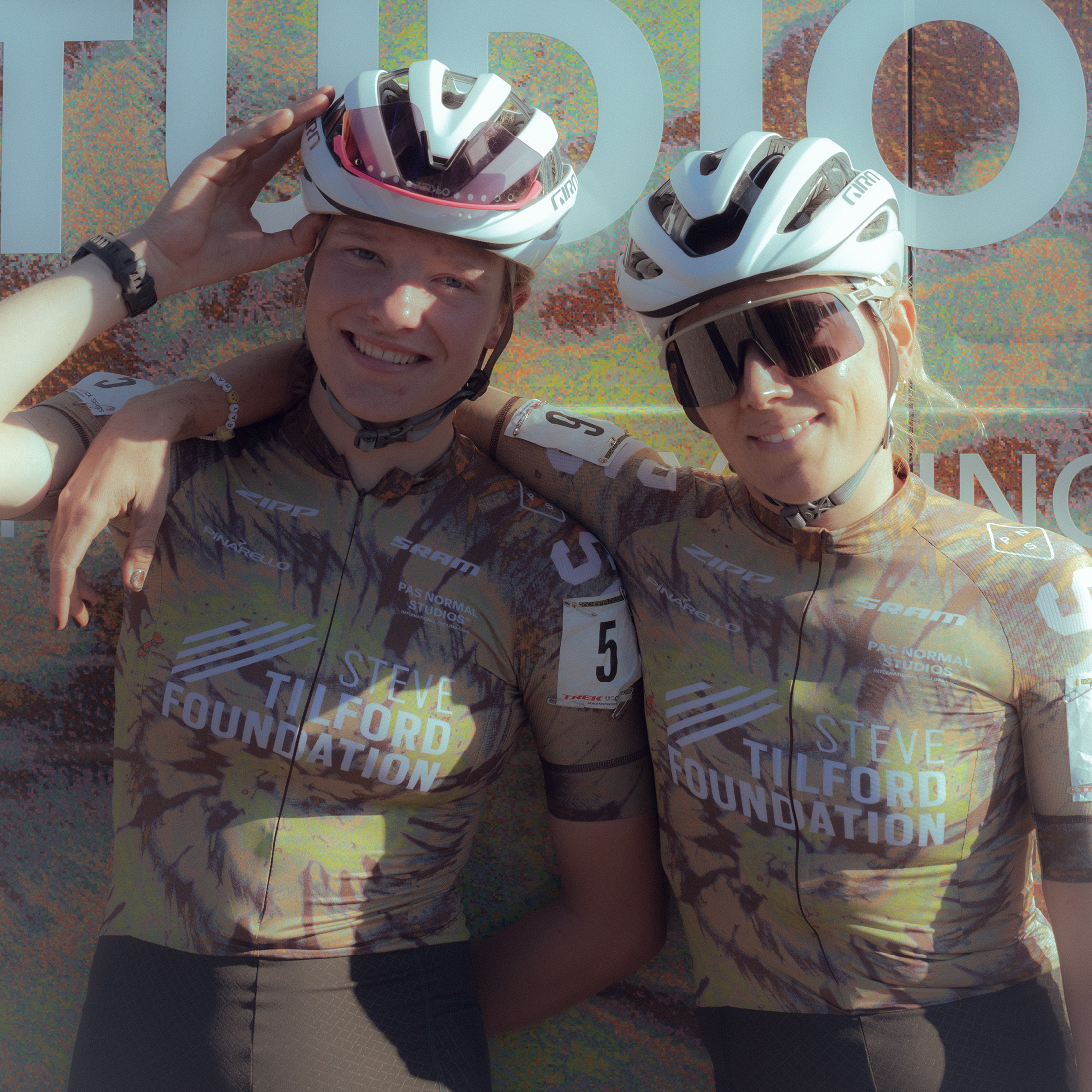 Two female cyclists wearing matching camouflage jerseys with white helmets celebrating at finish line
