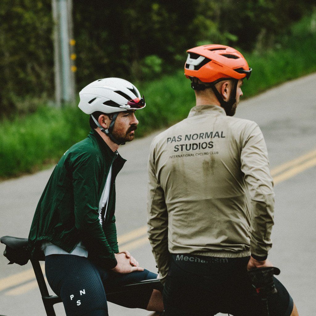 Two cyclists on road, one wearing green jersey, other in beige long sleeve with back branding