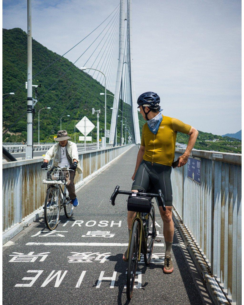 Cyclist wearing yellow jersey and grey shorts with time trial bike on bridge, local rider on city bike nearby
