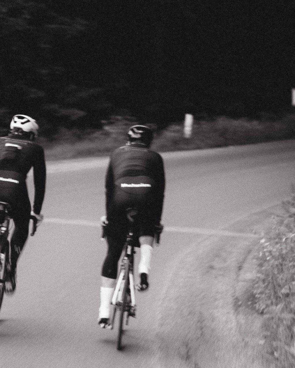 Two cyclists wearing dark jerseys with reflective details riding on road in black and white