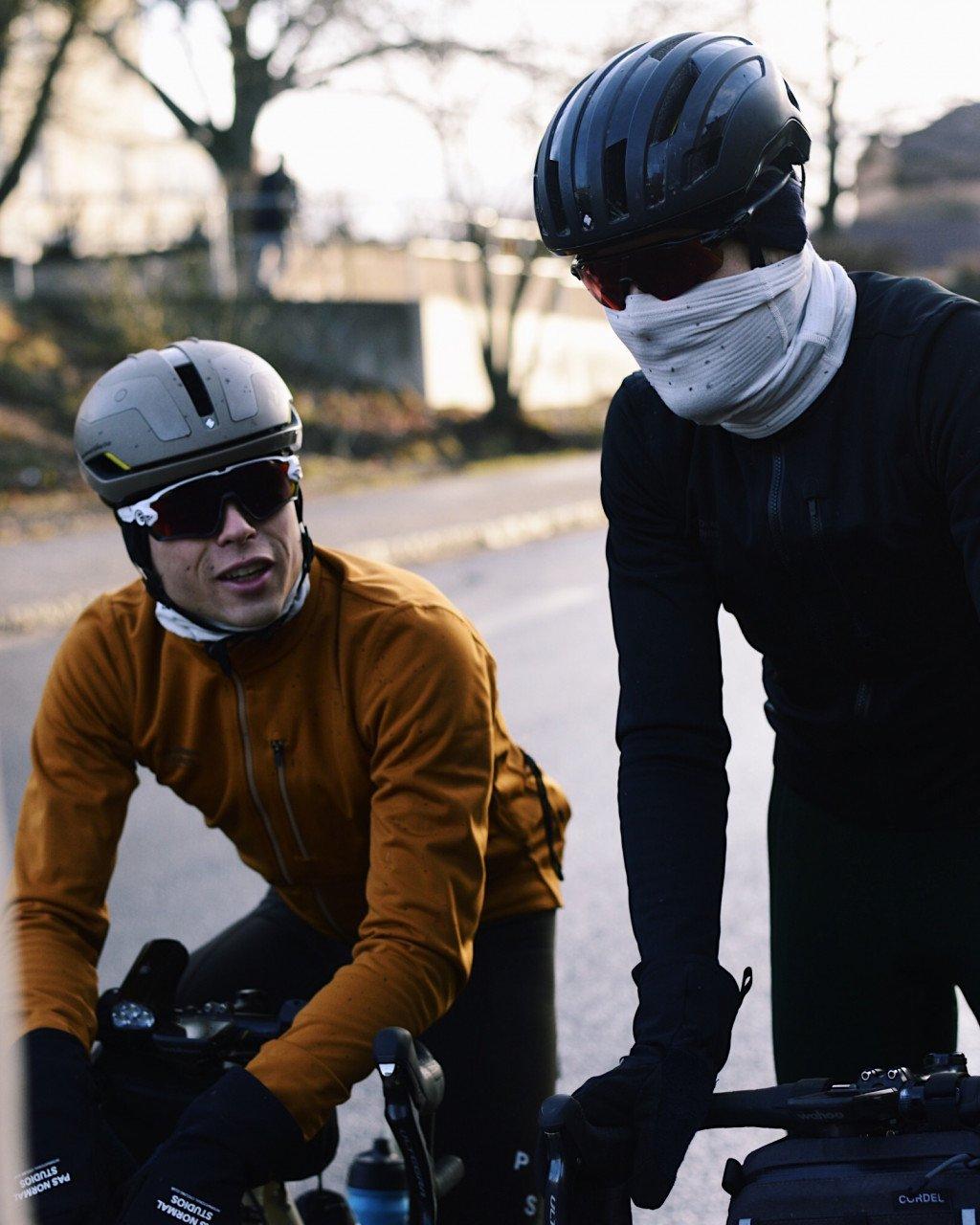 Two cyclists with aerodynamic helmets, one in orange jersey and one in black with white neck warmer, outdoors