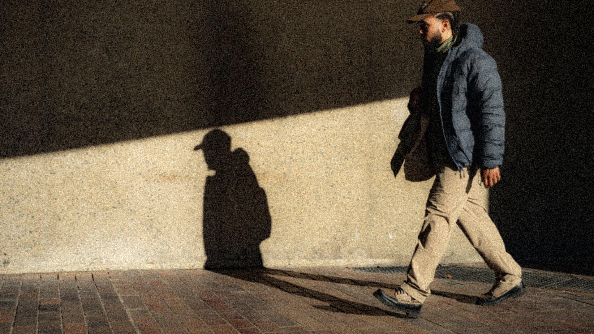 Model wearing navy quilted jacket and tan trousers walking on brick pavement with shadow cast on wall