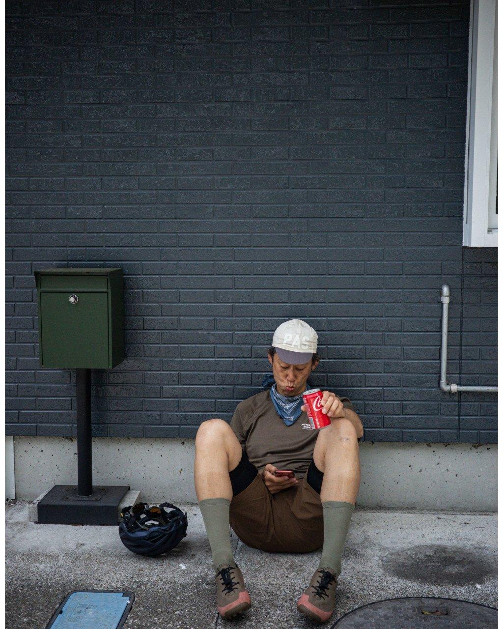 Model wearing brown cycling outfit and white cap taking a break, sitting against brick wall