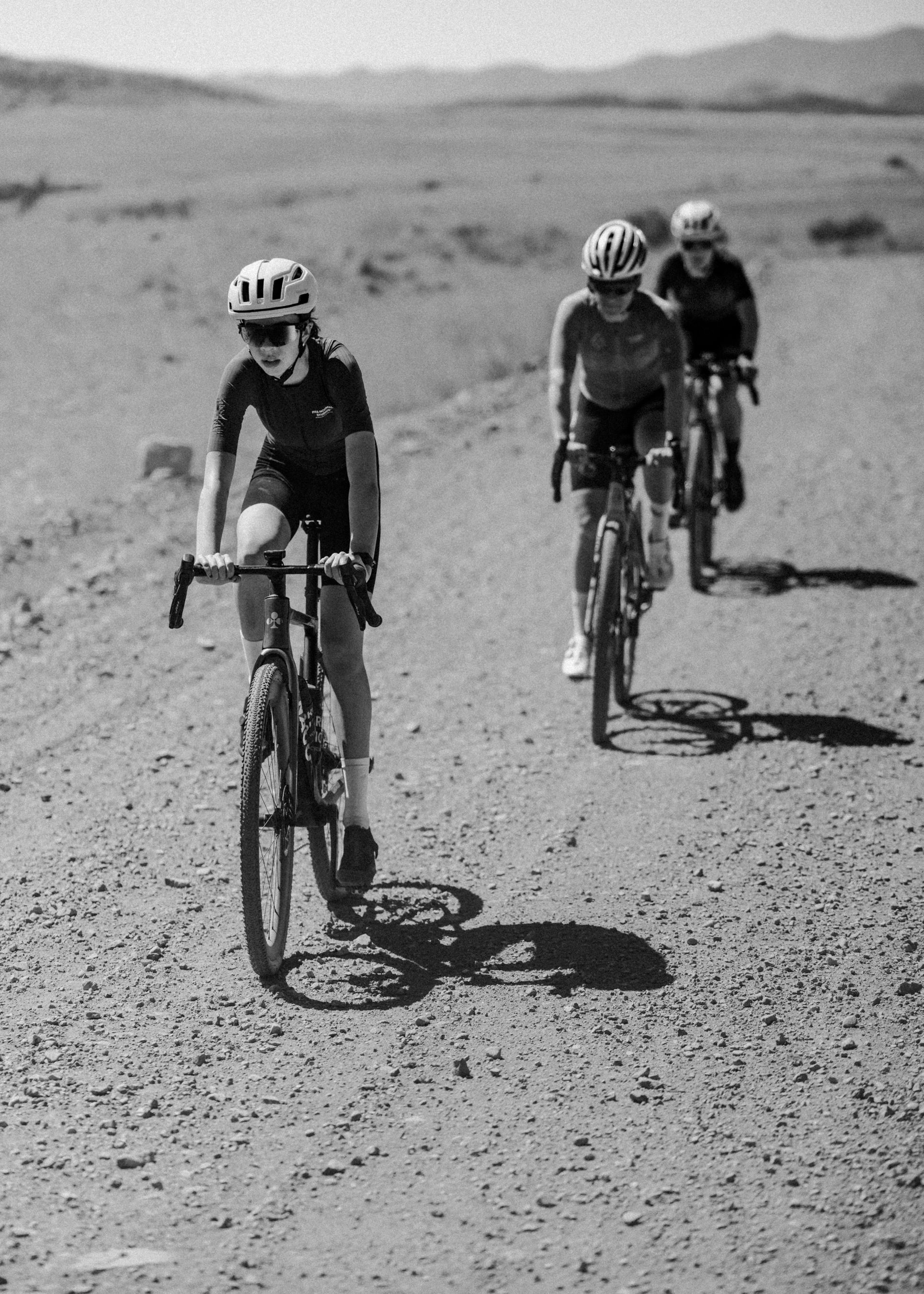 Three cyclists in time trial position riding gravel bikes across desert terrain in black and white