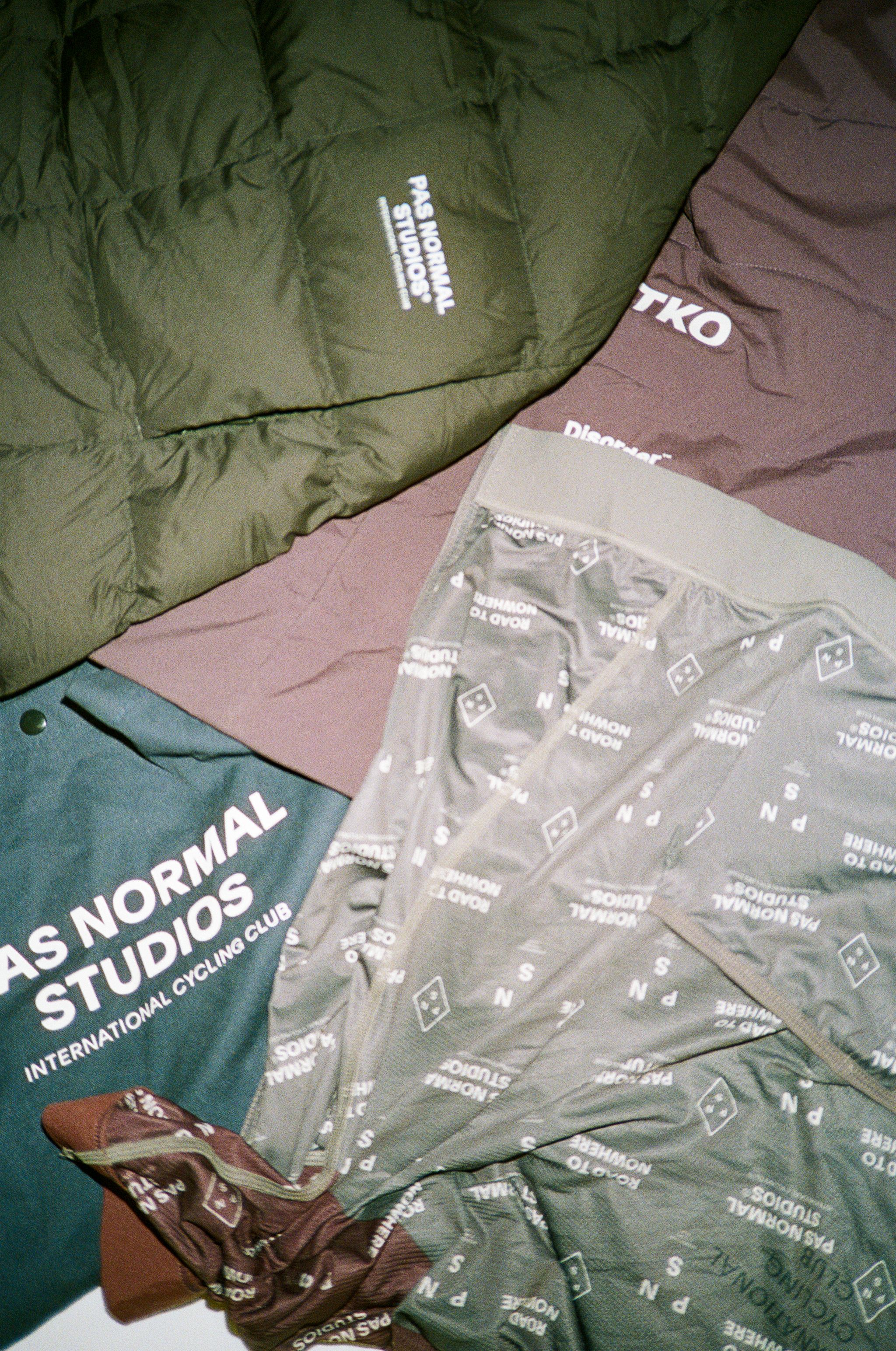 Flat lay of cycling apparel including green insulated jacket, blue jersey with white text, and patterned garments