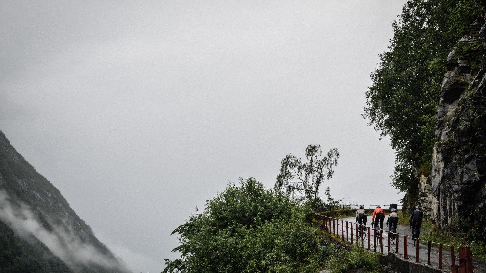 Cyclists on mountain road through misty Norwegian landscape with rocky cliffs and green vegetation