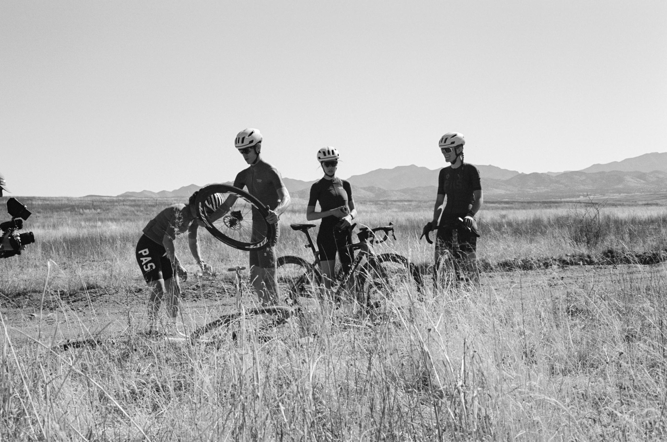 Models wearing black cycling apparel with bikes in desert grassland setting