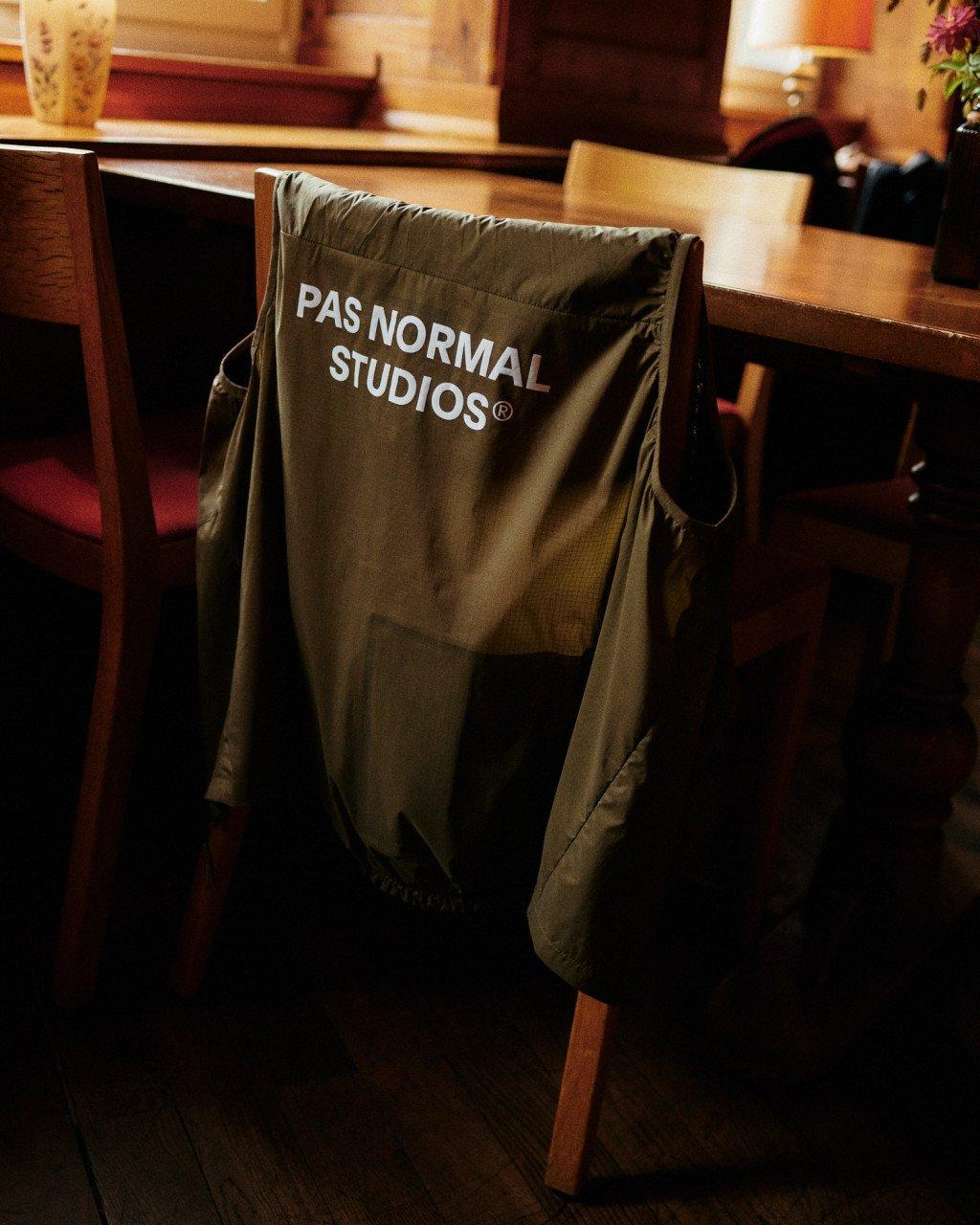 Olive green cycling jacket with white logo text draped over wooden chair in warm indoor setting