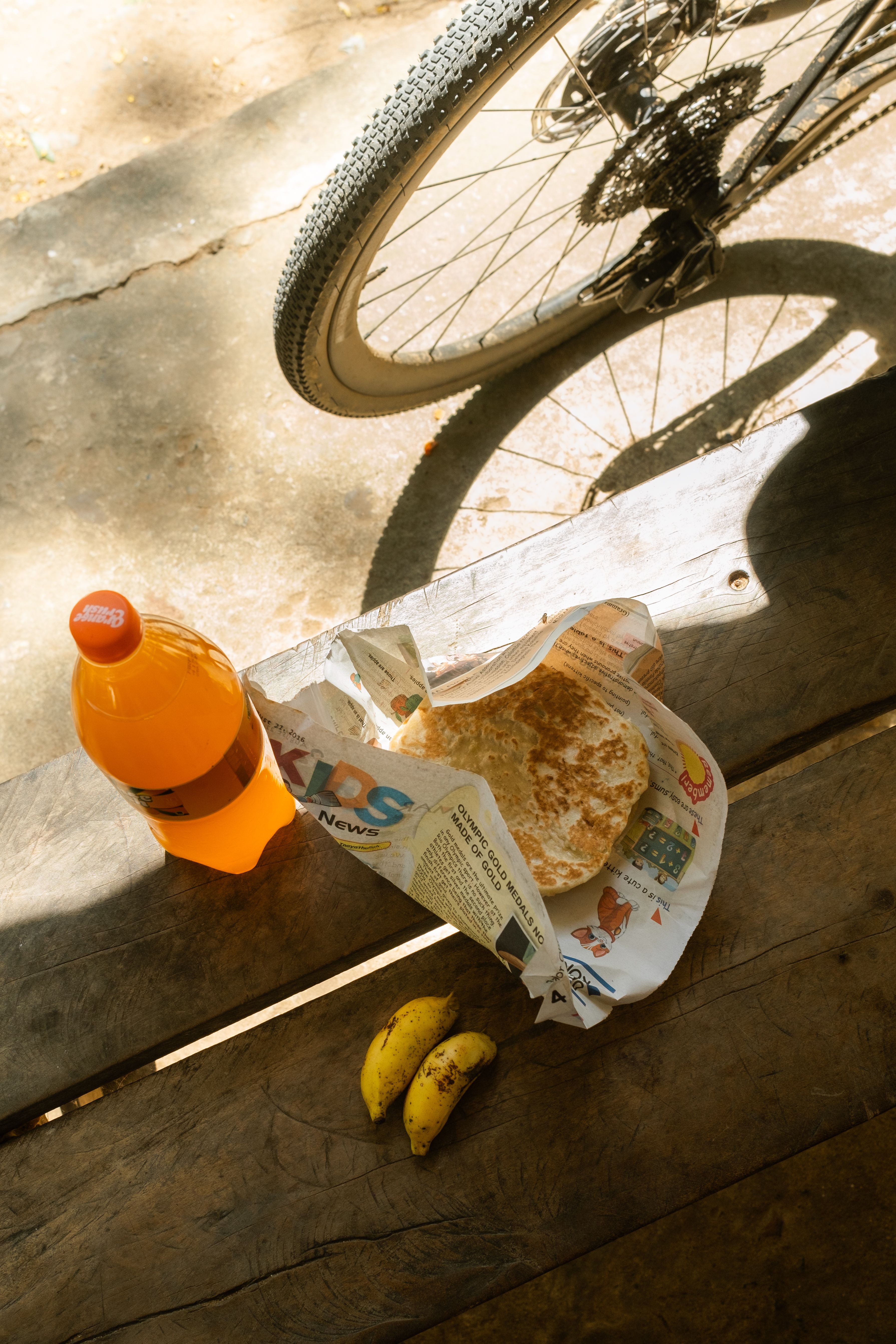 Cycling scene with bike wheel, orange water bottle, newspaper-wrapped food, and bananas on wooden surface