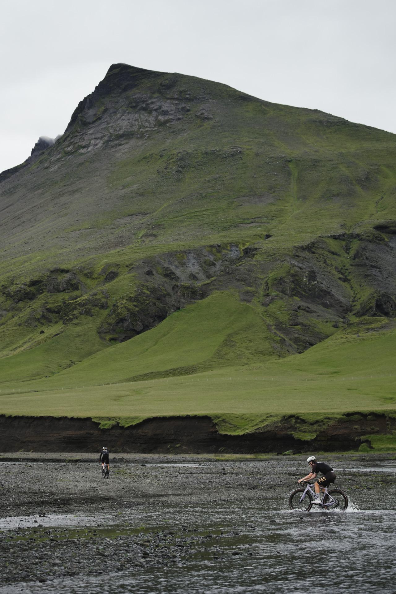 Cyclists riding across volcanic landscape with dramatic green mountain backdrop in Iceland