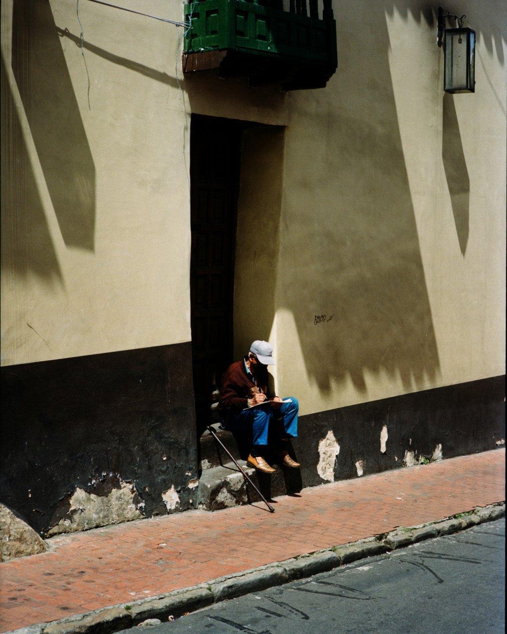 Person in white cap and dark red jacket sitting in shadowed doorway on urban street