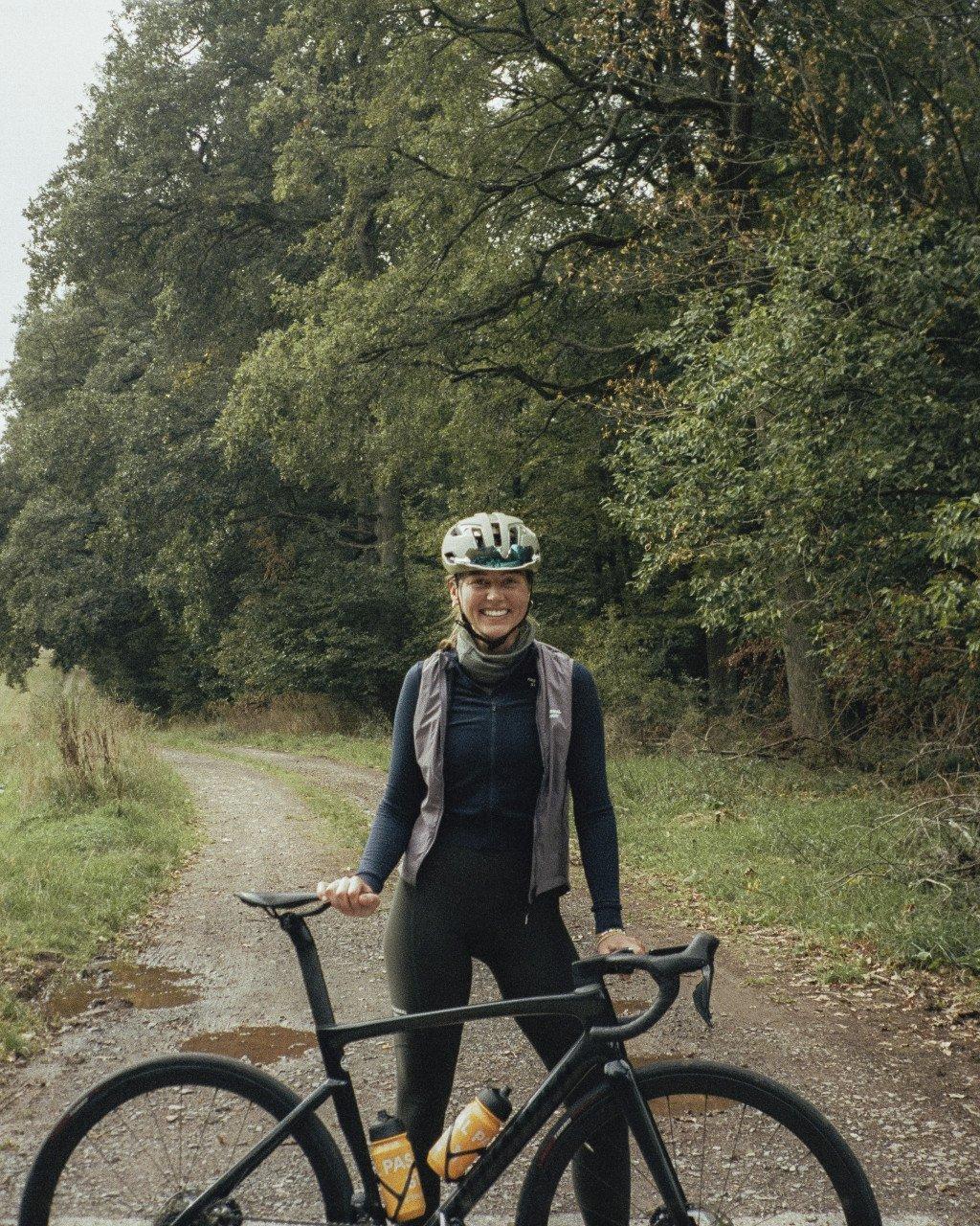 Model wearing black cycling outfit with grey vest and white helmet, standing with bike on country path