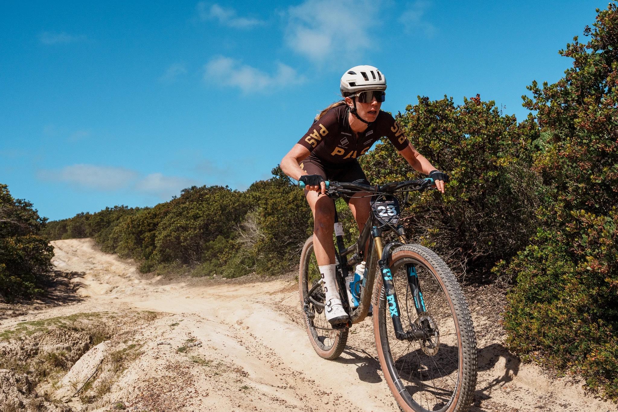 Model wearing brown cycling jersey with gold lettering mountain biking on dirt trail