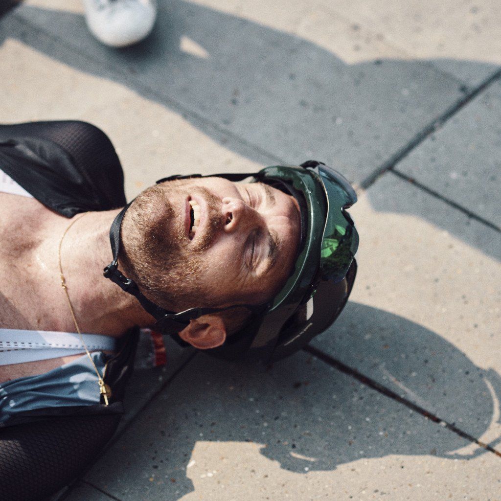 Model wearing green cycling cap lying on pavement with eyes closed in sunlight