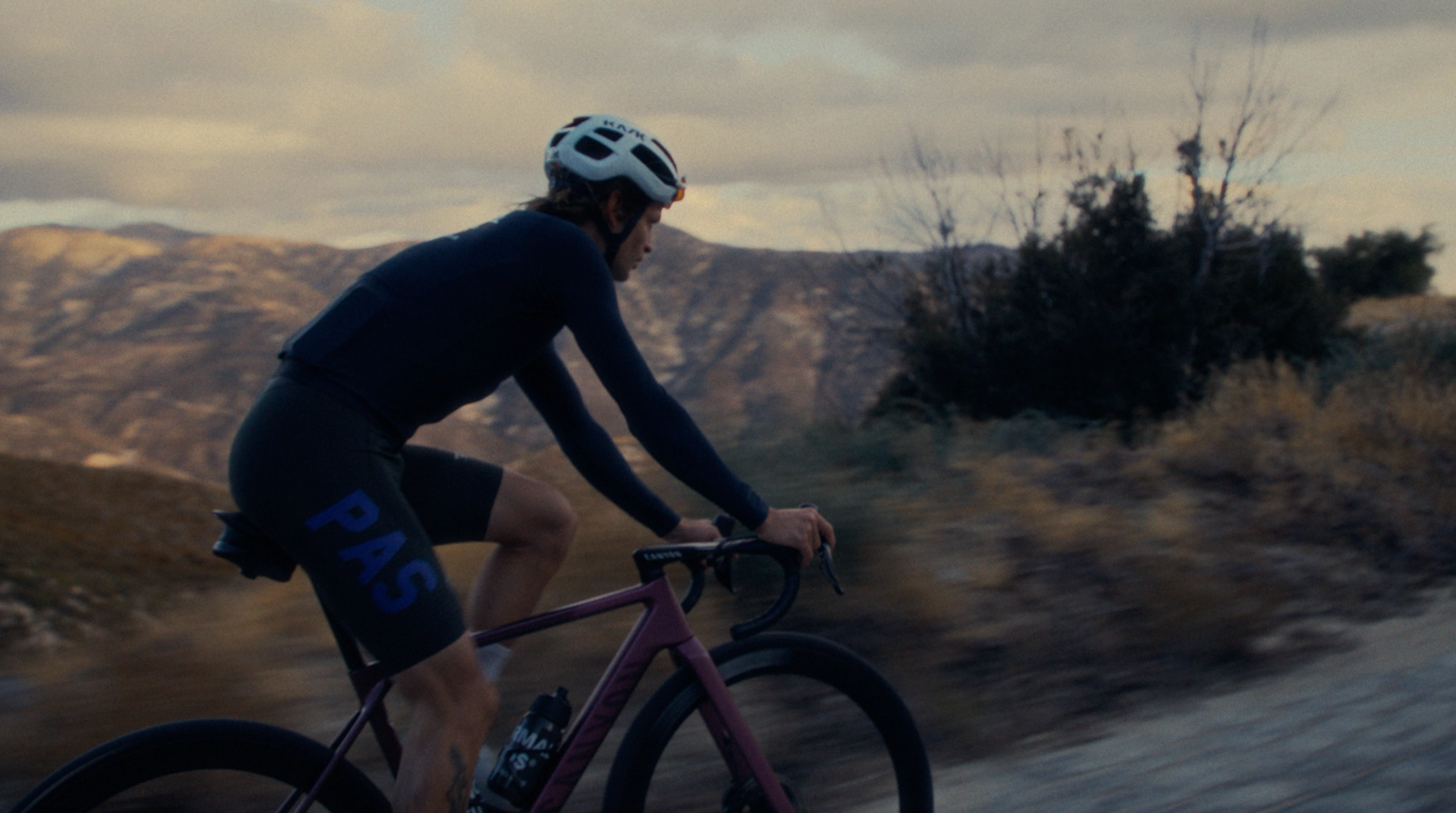Model wearing black cycling jersey and blue shorts on purple gravel bike in mountainous landscape at dusk