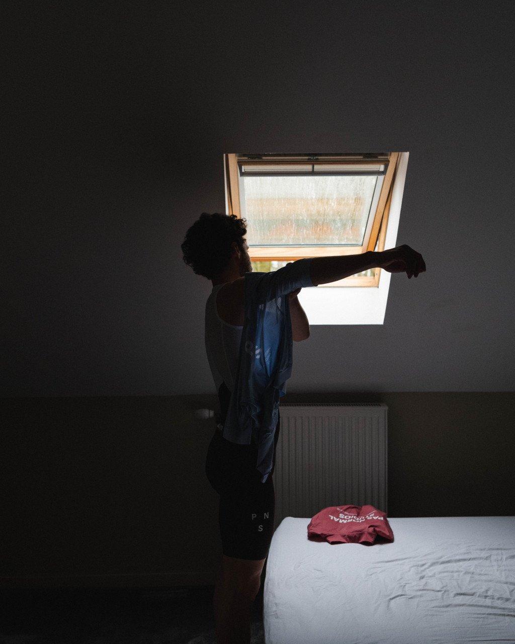 Model wearing dark blue cycling kit stretching by window with red jersey on bed