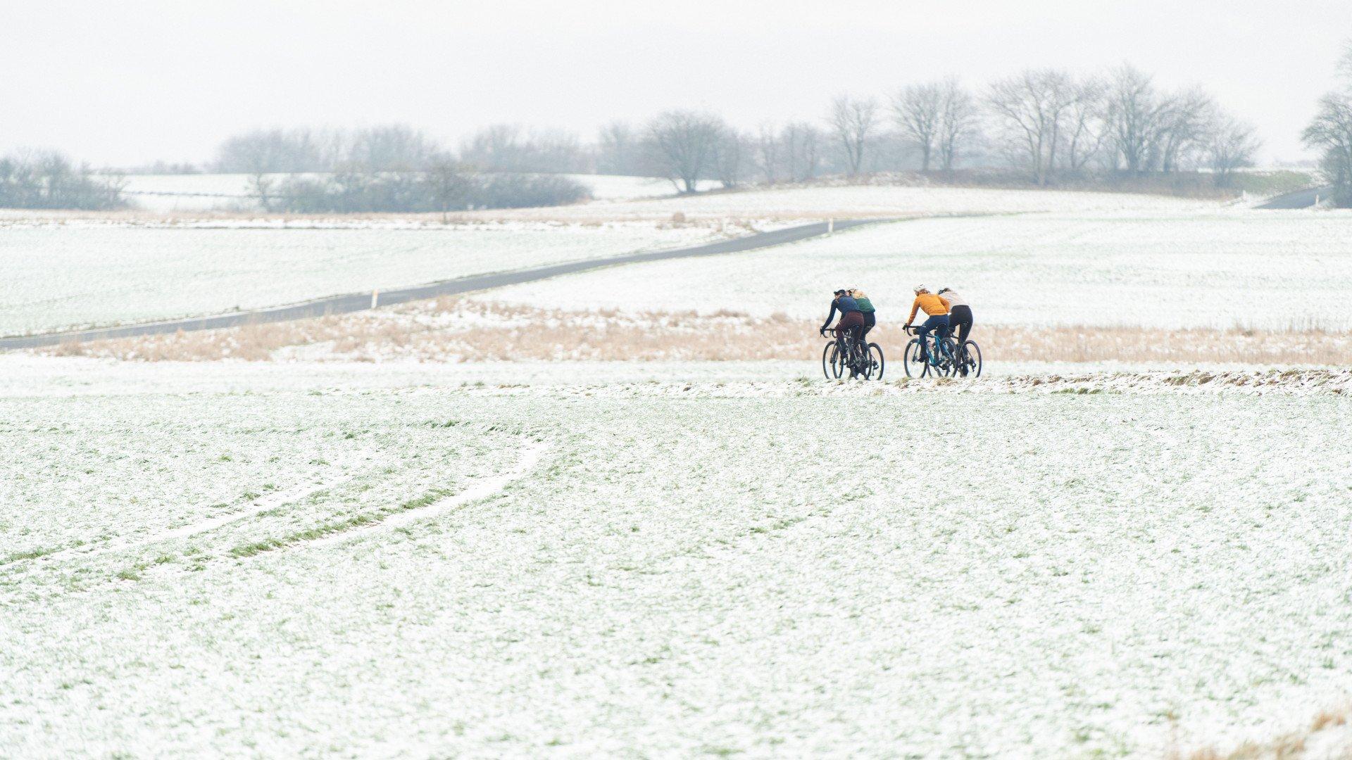 Two cyclists riding through snowy winter landscape on rural road