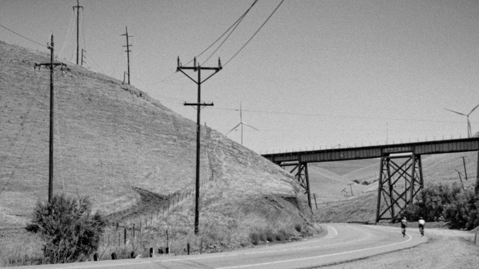 Black and white landscape with cyclists on desert road beneath railway bridge and power lines