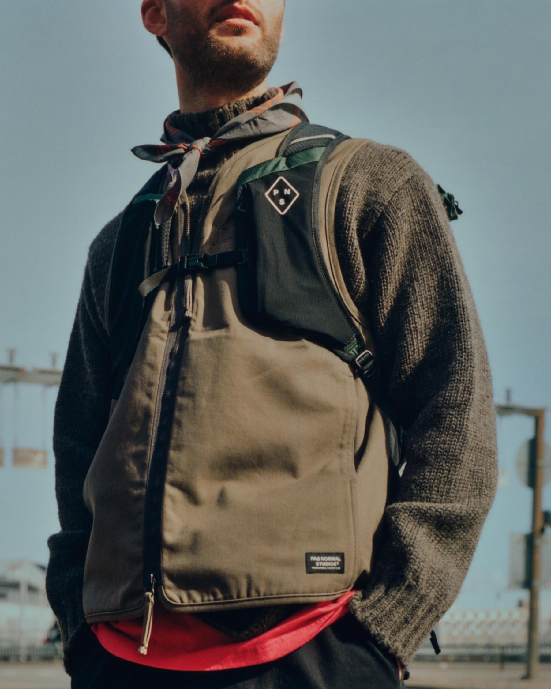 Model wearing beige backpack with diamond logo patch and adjustable straps in outdoor urban setting