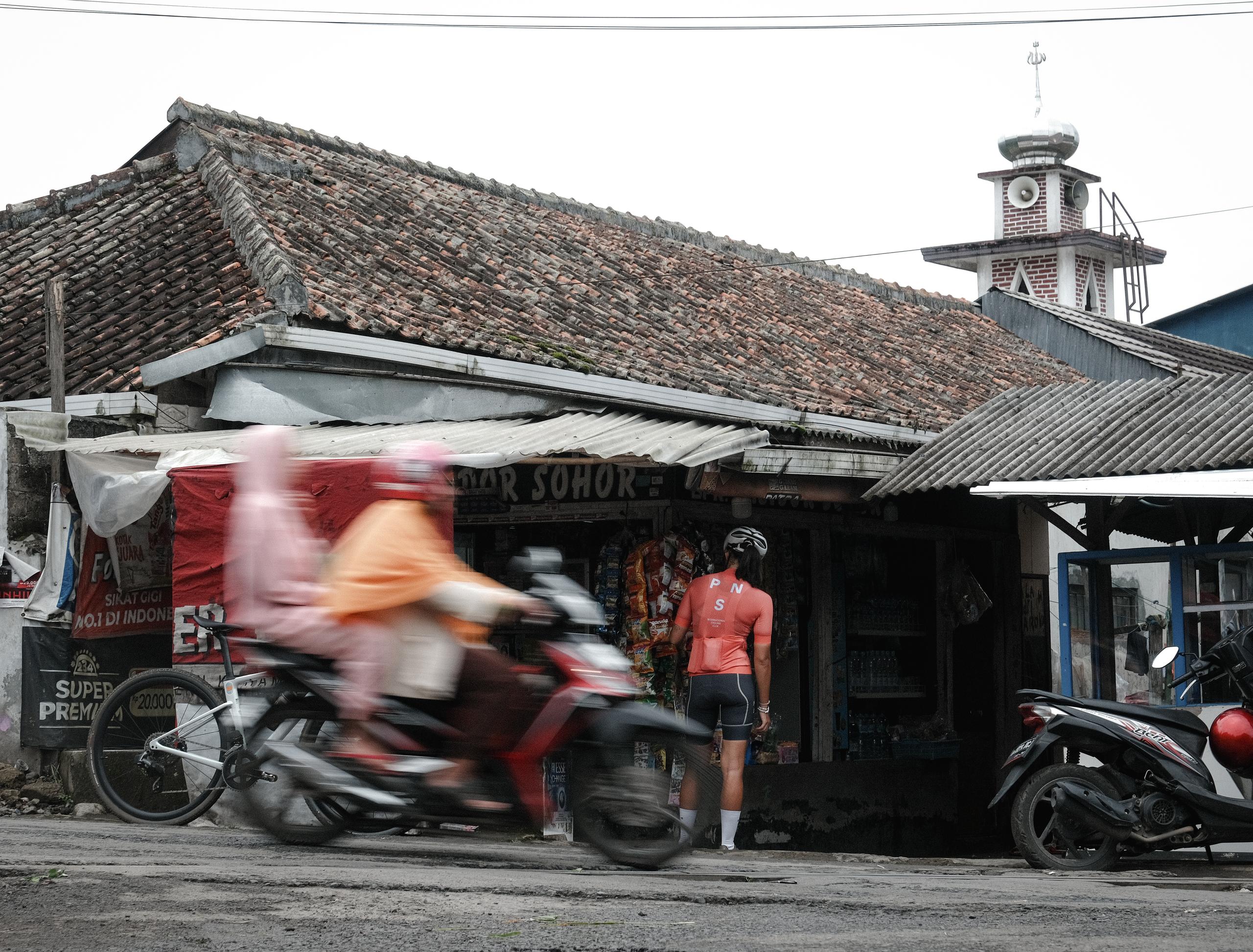 Model wearing red cycling jersey and black shorts standing outside Indonesian shop with motorcycle passing by