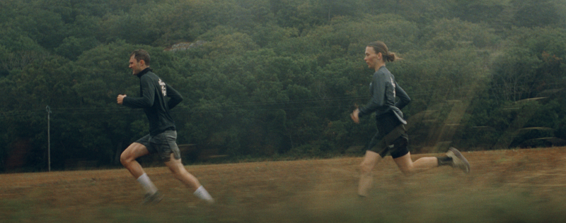 Two runners in grey athletic wear running on dirt track with forested hills in background