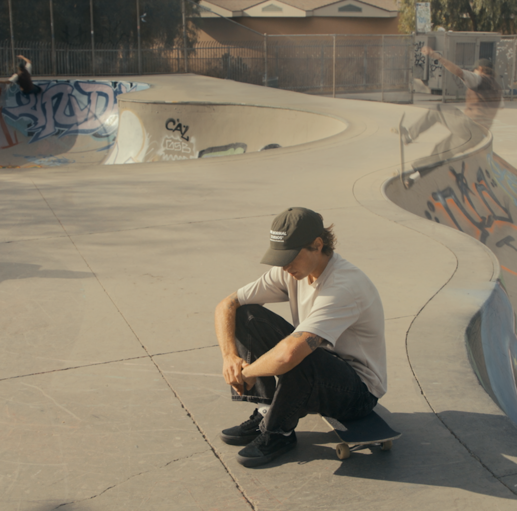 Model wearing white t-shirt and black cap sitting with skateboard at concrete skate park