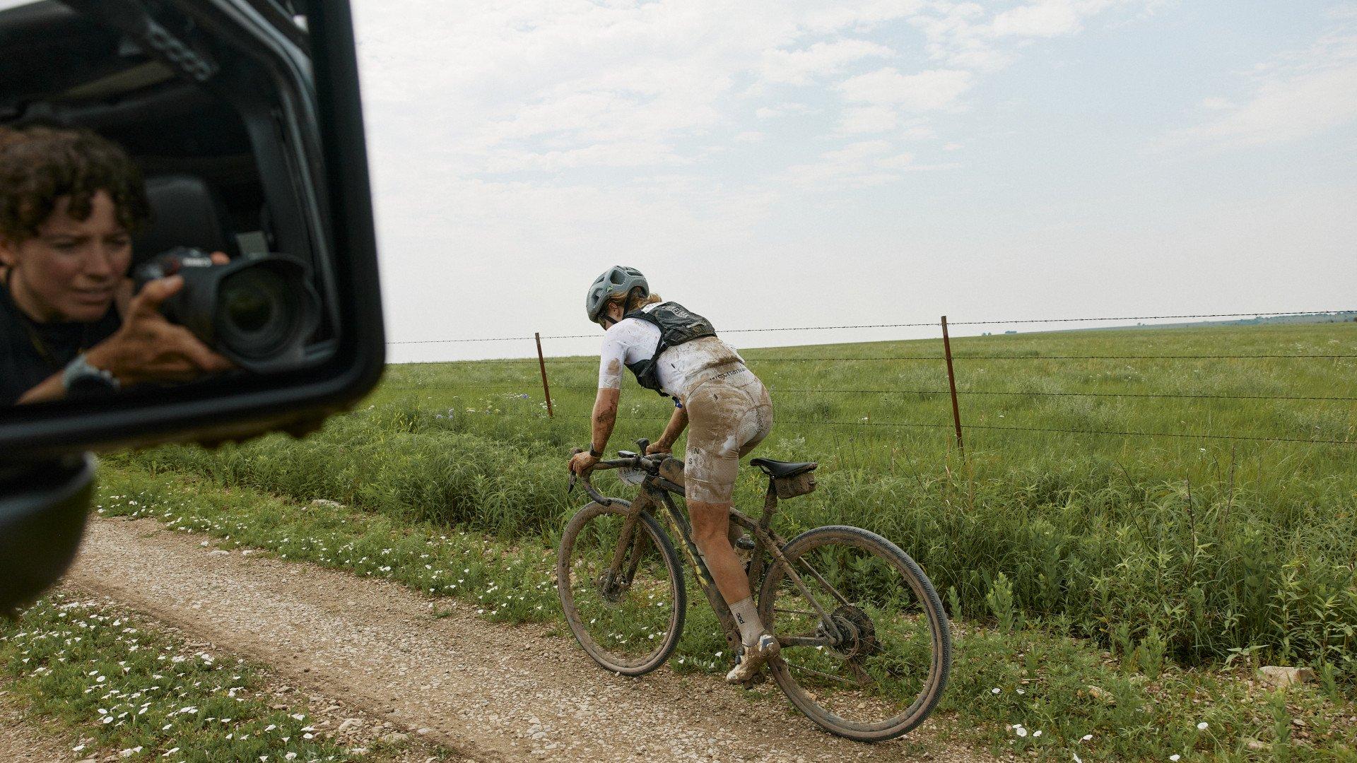 Cyclist on muddy gravel bike adjusting equipment roadside while photographer captures moment from vehicle
