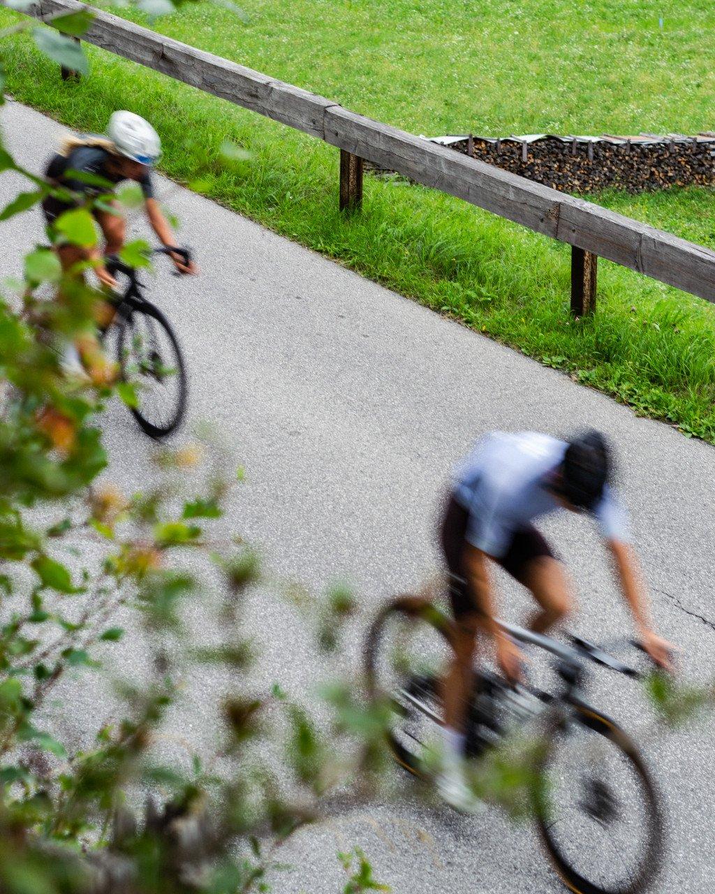 Two cyclists in motion blur racing along paved path beside wooden guardrail and green grass