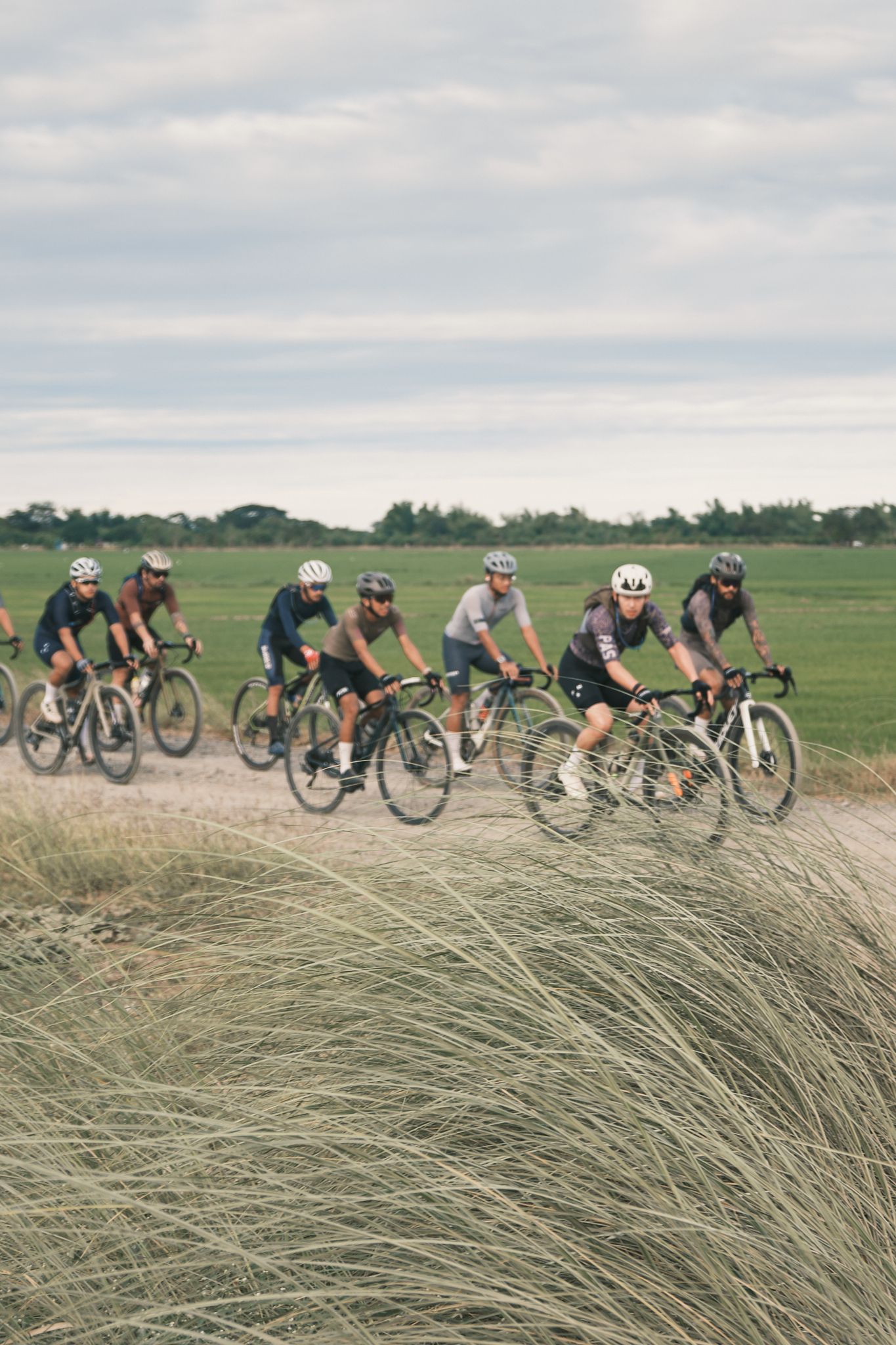 Group of cyclists riding gravel bikes on dirt path through countryside wearing mixed cycling apparel