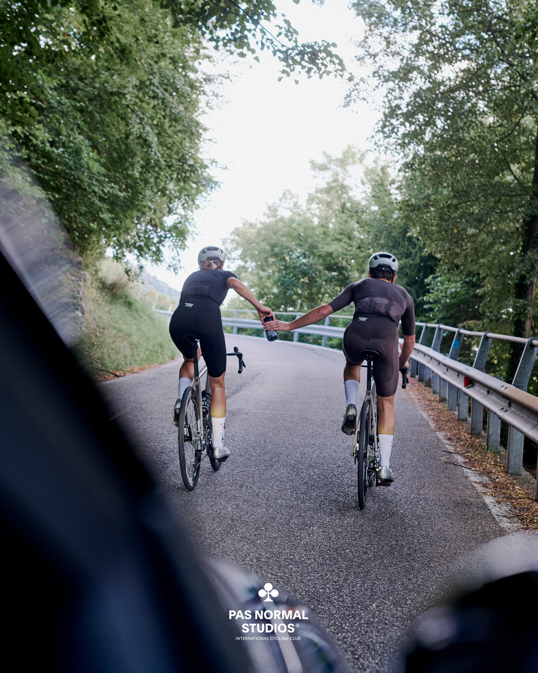 Two cyclists in black aerodynamic suits riding time trial bikes on country road, holding hands