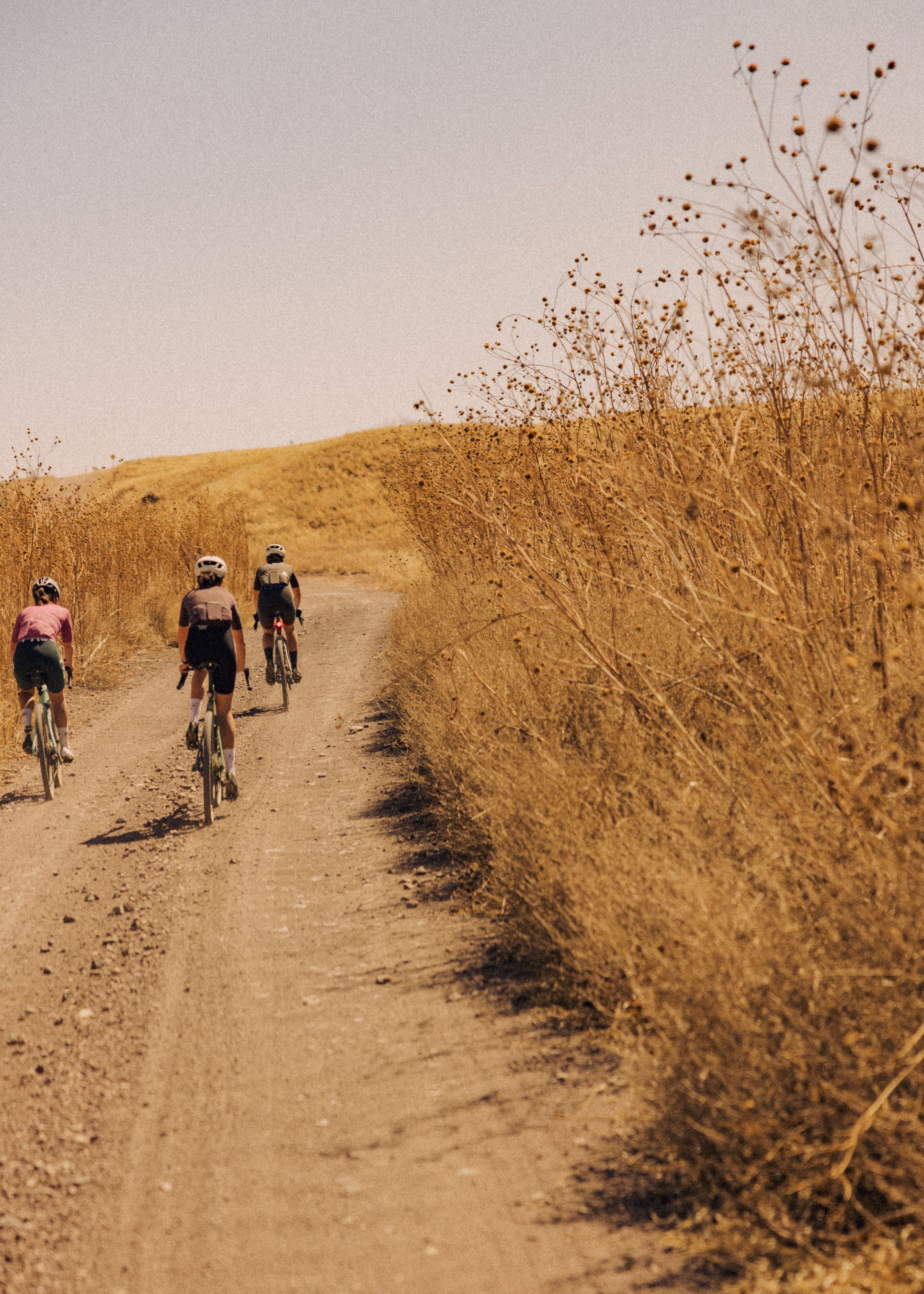 Cyclists riding gravel bikes on dirt road through golden hills with dry vegetation