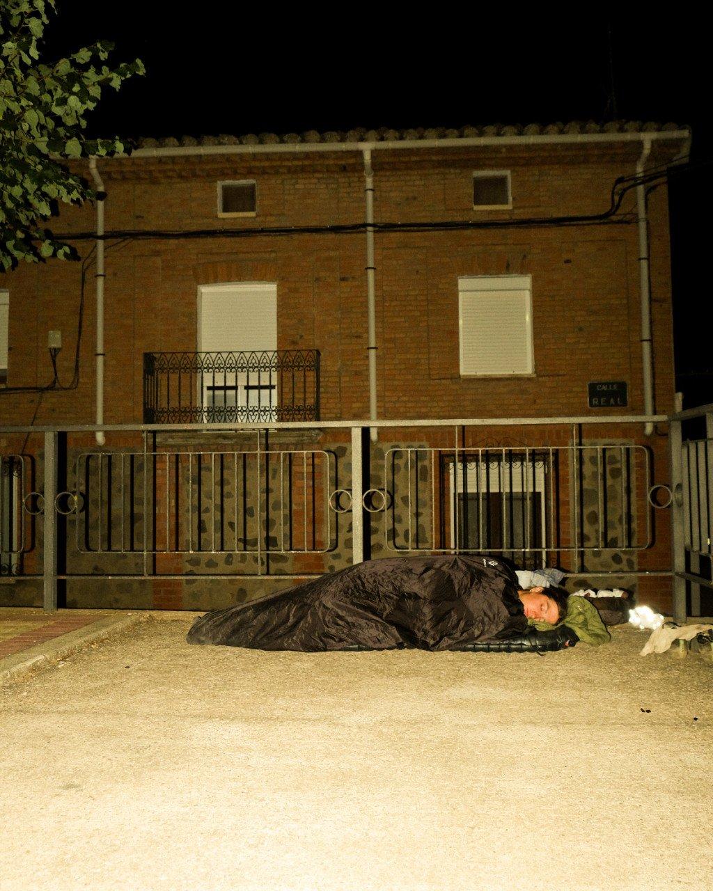 Person sleeping in black sleeping bag on street at night in front of brick building