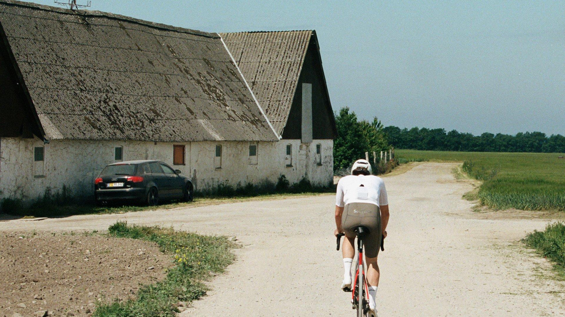 Cyclist in white jersey and beige shorts riding on rural gravel road past traditional farm buildings