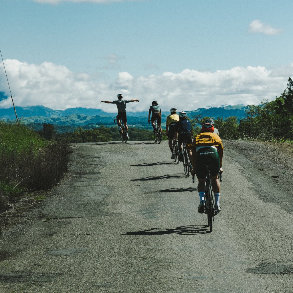 Group of cyclists on mountain road with one rider celebrating arms outstretched, mountainous landscape background