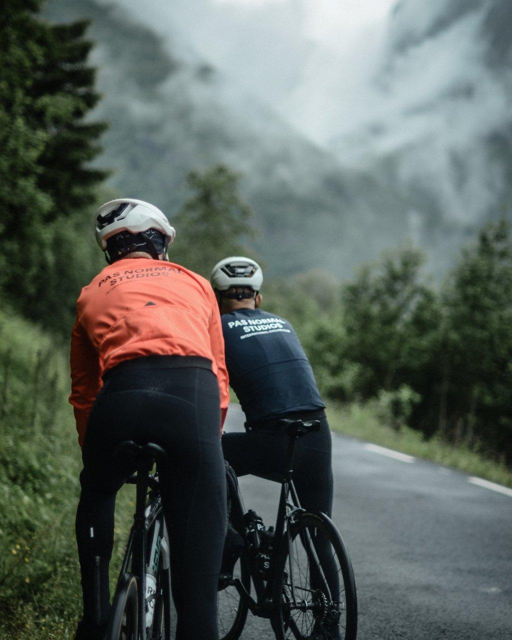 Two cyclists on mountain road wearing orange and navy jerseys with branded text
