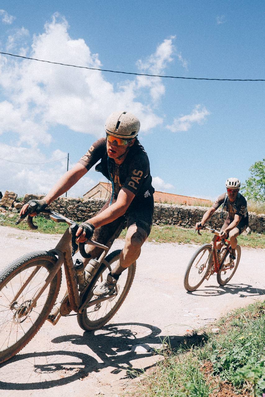 Models cycling on gravel road wearing dark cycling kits and helmets under blue sky with stone walls