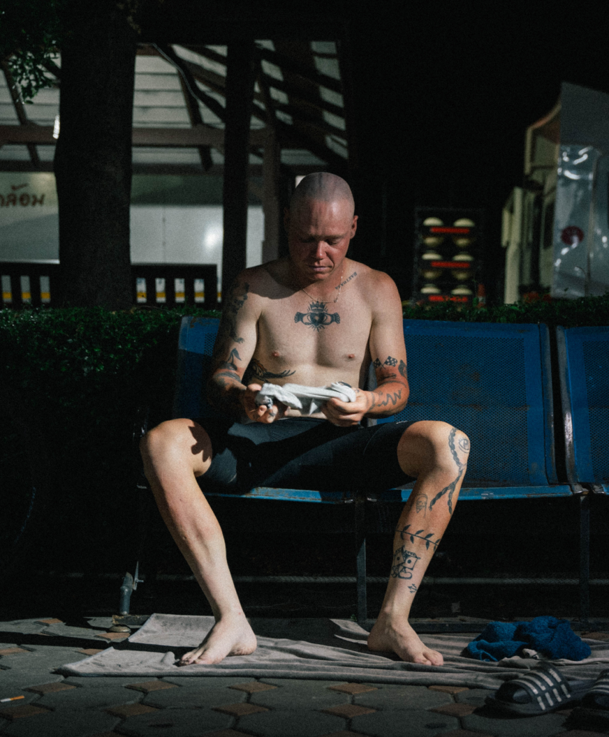 Model holding cycling jersey while sitting shirtless on blue bench in urban setting