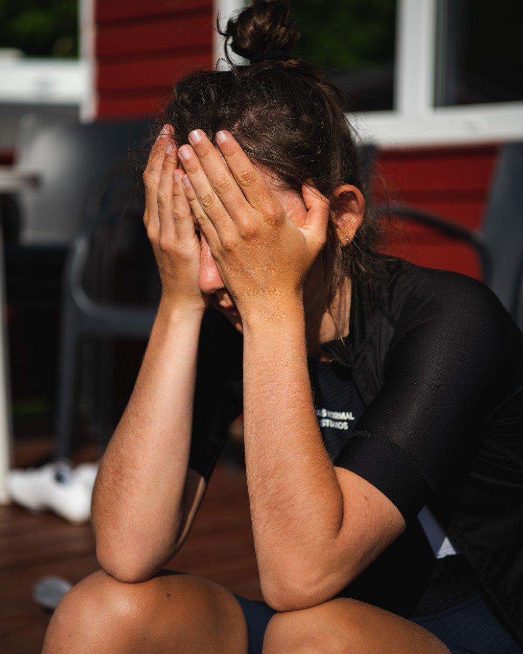 Model wearing black cycling jersey with hands covering face in indoor setting