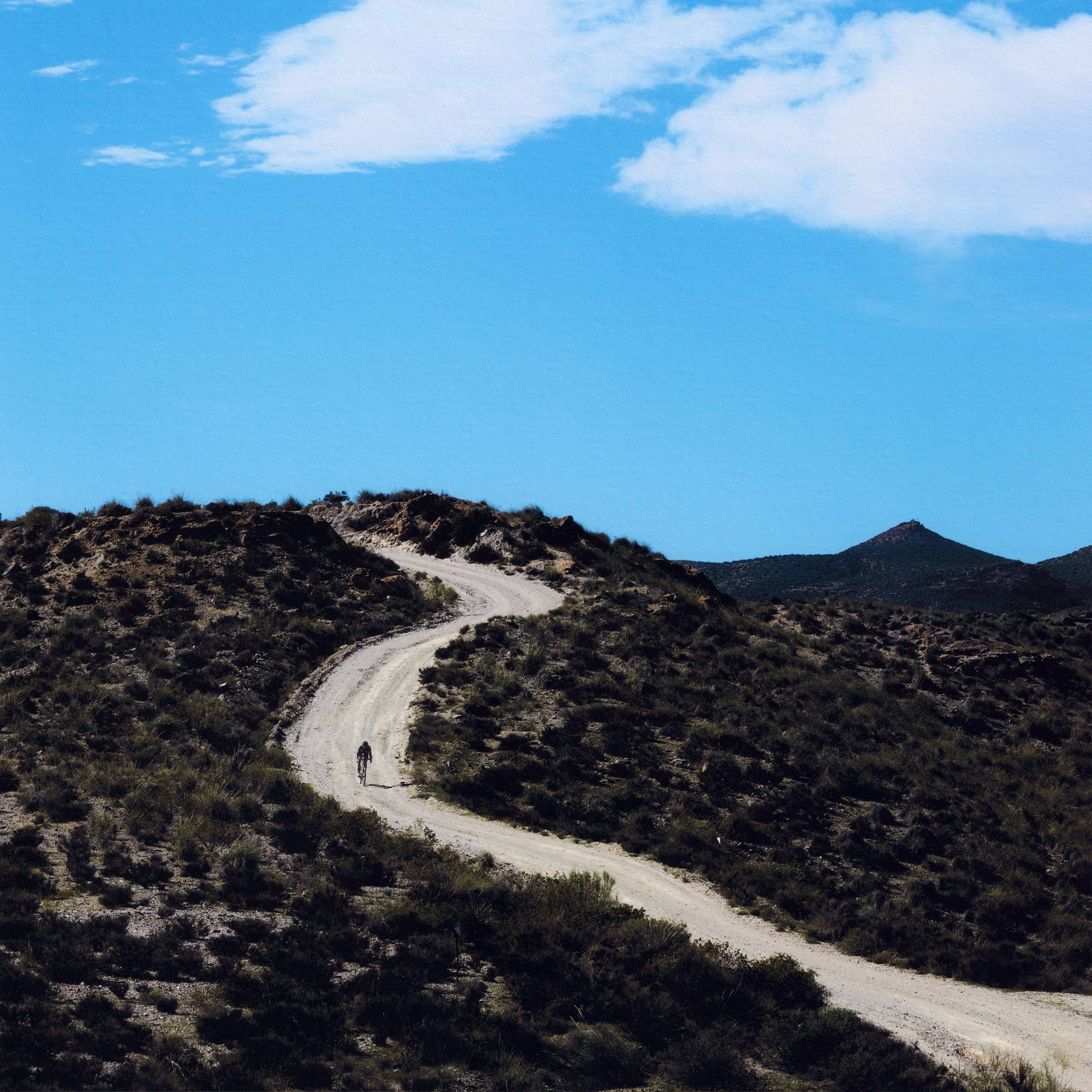 Cyclist on winding gravel road through desert hills under blue sky
