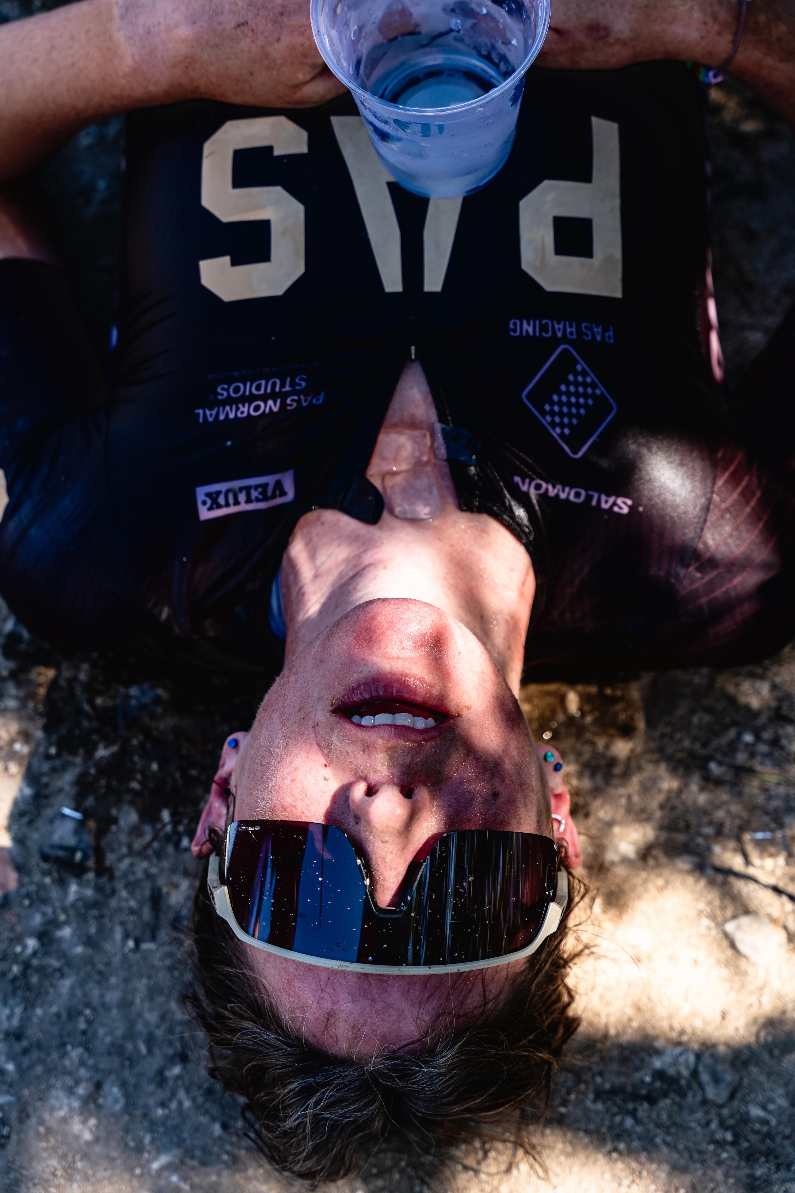 Model wearing black jersey with white lettering, lying on ground with reflective sunglasses and blue drink bottle