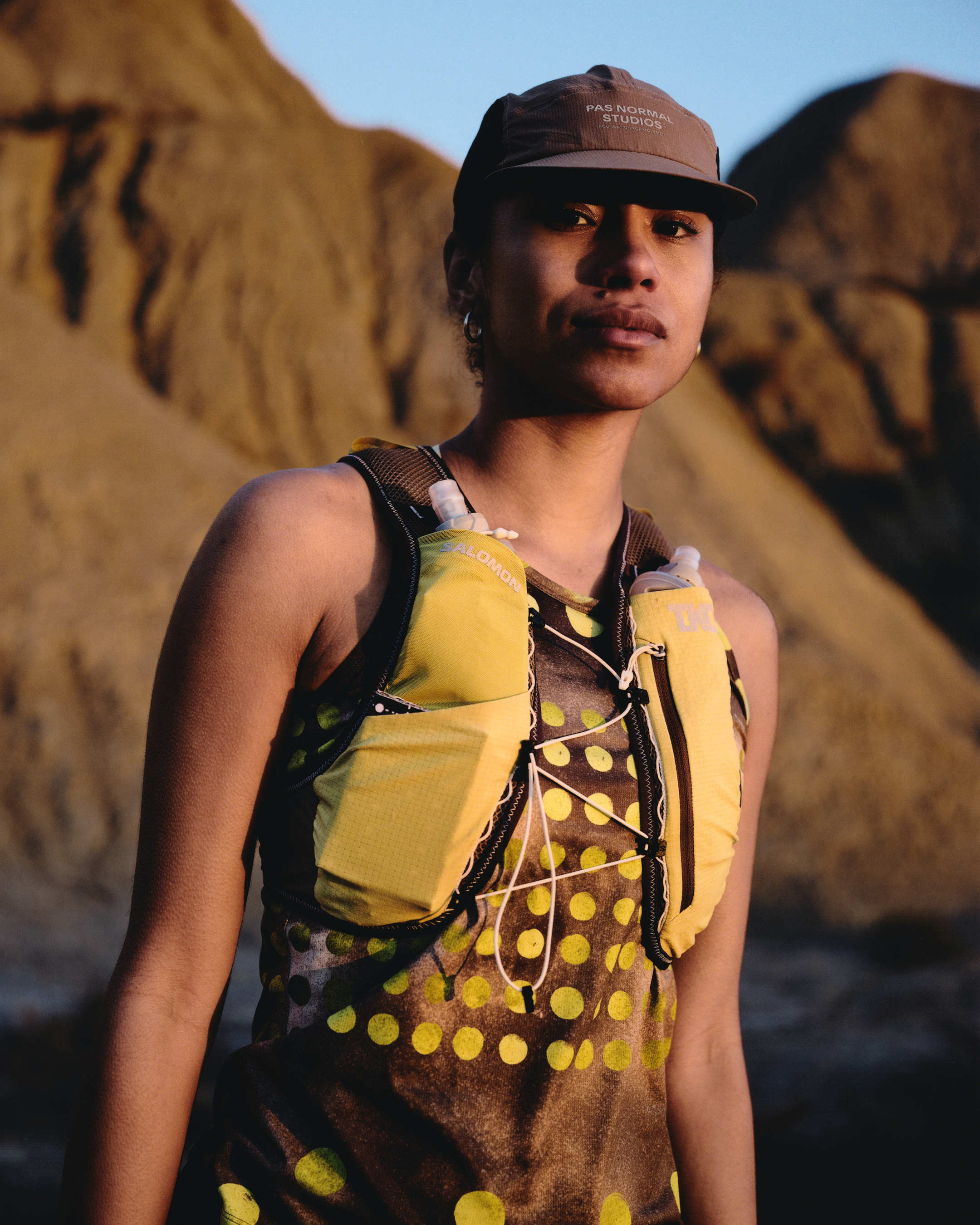 Model wearing brown cap and yellow polka dot running vest with hydration pack in desert landscape