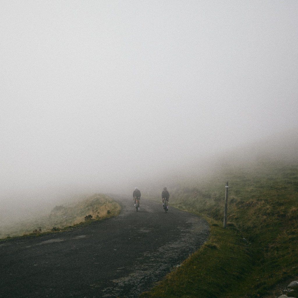 Two cyclists riding up misty mountain road through fog and moorland