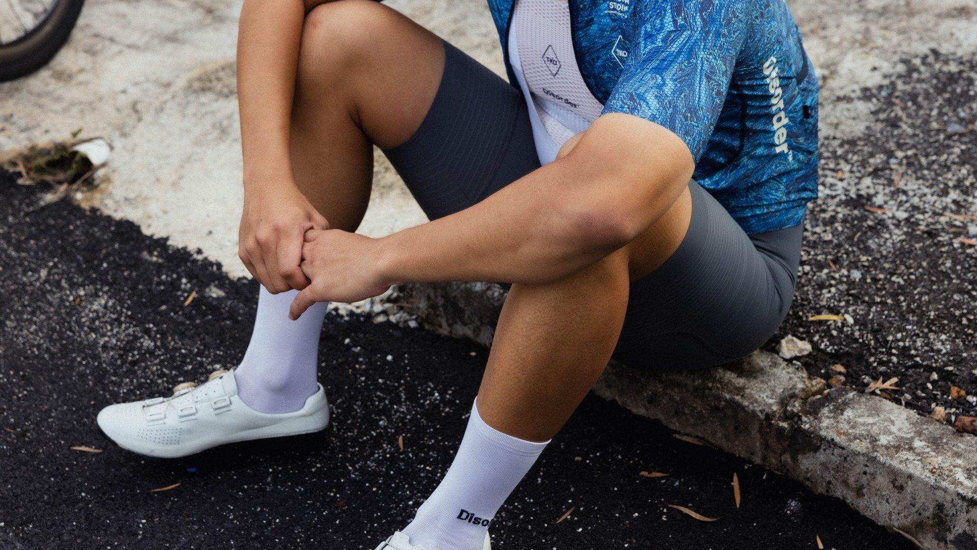 Model wearing blue patterned jersey with white socks and cycling shoes, seated on asphalt